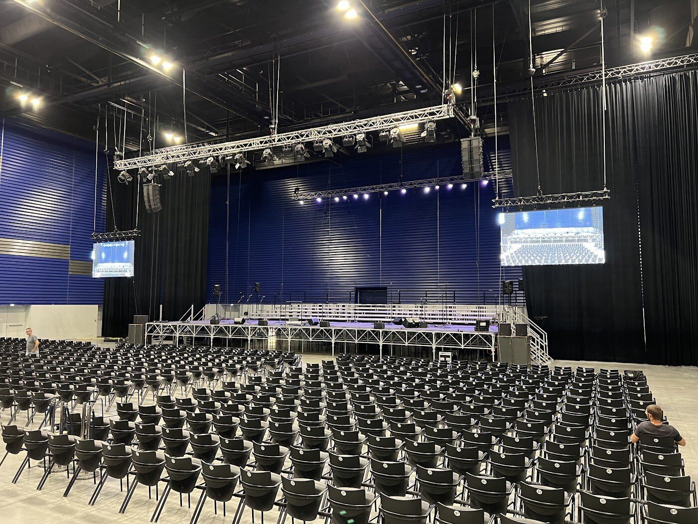 Grande salle en configuration assise : scène, diffusion accrochée en ligne source et deux écrans de renfort.